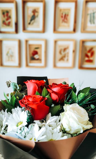 A floral arrangement featuring a bouquet of vibrant red roses with lush green leaves, complemented by white daisies and foliage, wrapped in soft pinkish-beige paper. The bouquet is positioned on a tab