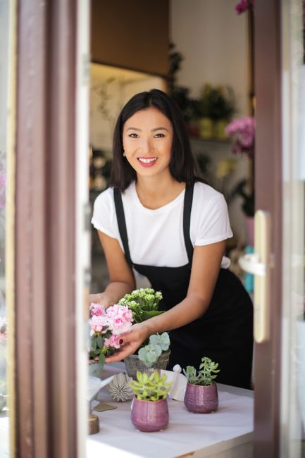 A smiling young woman with shoulder-length black hair, wearing a white t-shirt and a black apron, is standing inside a floral shop, arranging a bouquet of fresh pink and white flowers with green folia