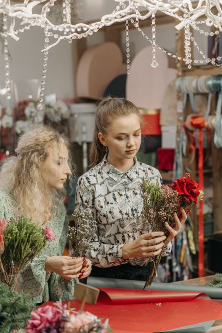 A young woman with her hair tied back, wearing a patterned blouse, holding a small bouquet of red roses and mixed greenery, stands indoors in a florist workshop. An older woman with curly blonde hair 