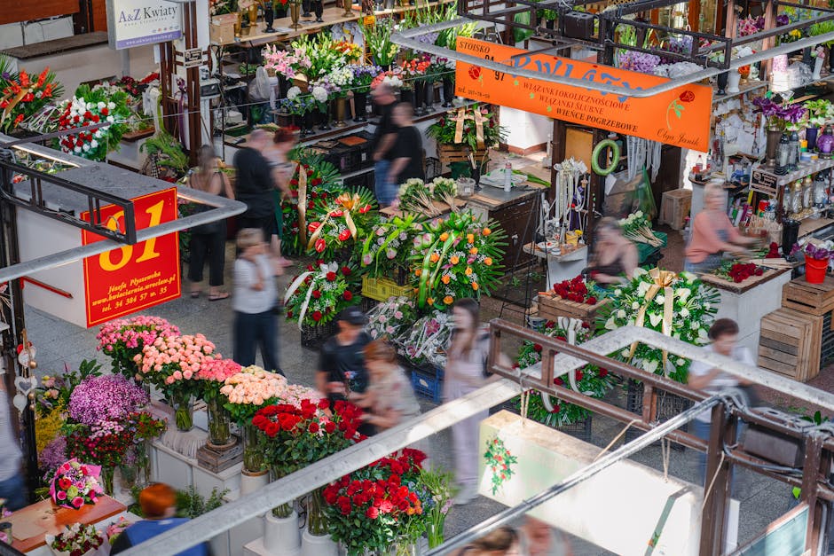 A busy indoor flower market showcasing a vibrant array of floral arrangements and bouquets, including pink, red, purple, and white blooms such as roses, chrysanthemums, and lilies, displayed on tables