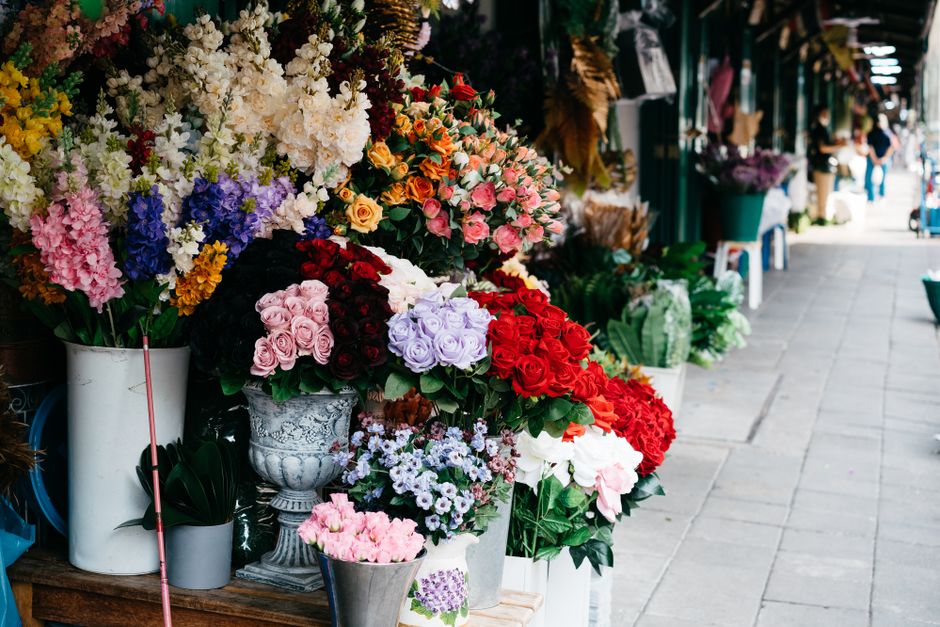 A vibrant display of fresh floral arrangements arranged in white ceramic and metallic vases on an outdoor flower shop stall, featuring a variety of roses, hydrangeas, and other blooms in shades of pin