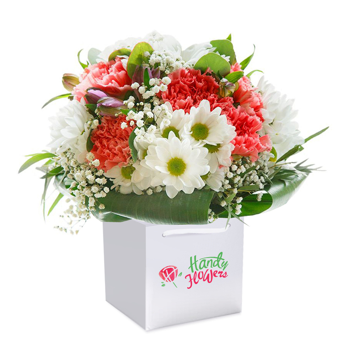 An exuberant, meticulously arranged bouquet finds its place in a spotless square white gift bag, effortlessly set against a seamless white background-the hallmark of Osidge's thoughtful yet contemporary floristry. Rounded and full, the bouquet features a striking combination of carnation blossoms-ranging from tender peach to vibrant coral and deep red-each bloom defined by intricately ruffled petals that promise both visual and tactile delight. These are skillfully intermingled with pristine white daisy-like flowers, their chartreuse centers offering an invigorating contrast and a dash of Osidge cheer. Wispy bouquets of baby's breath are spread throughout, their airy clusters creating a cloud-like texture ideal for adding volume without heaviness. Subtle purple-hued buds emerge from one side, adding intrigue and the understated beauty of anticipation familiar to anyone strolling the quiet avenues of Osidge. At the base, a border of rich, glossy green foliage and lush, broad leaves echoes the local landscape, creating a supportive frame that unites the arrangement. The gift bag's white ribbon handles suggest an easy handoff-perhaps for same-day flower delivery to a birthday celebrant or to brighten a home in the Osidge area. Each floral element has been purposefully chosen and arranged, a testament to the experience and love of local Osidge florists dedicated to quality and customer delight on every occasion.