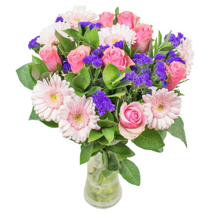 A luminous floral bouquet rests within a clear glass vase, its composition a testament to the natural beauty found throughout Osidge. In the heart of the arrangement, seven to nine roses bloom in varying pinks, their petals at different stages of opening, each spiral a nod to classic elegance. Interlaced with these are five to seven large-headed Gerbera daisies, their blush-pink petals radiating outwards and nearly white at the centres, much like the wildflowers that accent the edges of Brunswick Park in spring. Pops of vibrant purple statice weave through the arrangement, giving depth and a playful touch of contrast that makes the pinks feel even more radiant. Abundant, deep green foliage fans from the base, evoking the lushness along Waterfall Walk, while the sturdy clear vase grounds the whole display in simple sophistication. Every stem is expertly placed to highlight its individuality yet contribute to the overall sense of fullness and delight. Suitable for birthdays, Mother's Day, or just to brighten a favourite nook in any Osidge home, this bouquet brings together tradition and freshness-reflecting the warm character of the neighbourhood, with every petal chosen by local florists proud to serve their community.